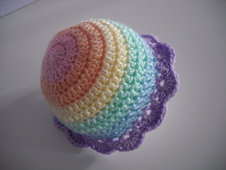 Puede incluir: Un gorro de ganchillo con un patr&oacute;n de degradado de arco&iacute;ris en tonos pastel de hilo rosa, naranja, amarillo, verde y azul. El gorro est&aacute; rematado con un borde festoneado de color morado, creando un dise&ntilde;o circular texturizado con puntos visibles.