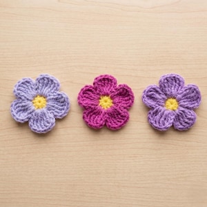 May include: Three crocheted flower appliqués arranged in a horizontal row. The left flower is light purple, the centre flower is magenta, and the right flower is a medium shade of purple. Each flower has five rounded petals and a yellow centre.