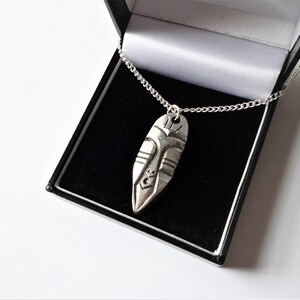 May include: A silver necklace with a pendant shaped like a stylized face. The pendant is detailed with geometric patterns and is hanging in a black velvet box.