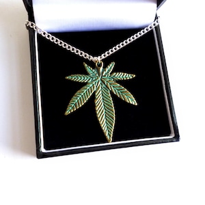 Könnte beinhalten: Eine silberne Kettenkette mit einem grünen und goldenen Marihuana-Blatt-Anhänger. Der Anhänger befindet sich in einer schwarzen Samtbox.