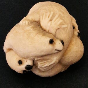 Op de afbeelding: Een gebeeldhouwde sculptuur van drie zeehonden die in elkaar verstrengeld zijn. De zeehonden hebben een crèmekleurige beige kleur met zwarte ogen en zijn gebeeldhouwd in een afgeronde, compacte vorm. De sculptuur staat tegen een donkere achtergrond.