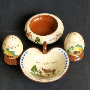 May include: A vintage ceramic kitchen set featuring a small apple-shaped dish, a round dish, and two egg-shaped salt and pepper shakers. The set is cream-colored with brown accents and hand-painted illustrations of houses and trees. The apple dish has the text "Knaresborough".