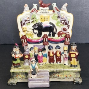 Könnte beinhalten: Eine Keramikfigur einer Zirkusszene mit dem Titel "Wombwell's Menagerie", die einen schwarzen Elefanten, verschiedene Tiere und eine Gruppe kleiner Figuren auf einer Plattform mit Stufen zeigt, bemalt in Farben wie Rot, Gelb und Grün.