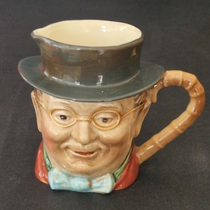Könnte beinhalten: Keramikkrug in Form eines lächelnden Mannes mit Zylinder und Brille. Der Griff ist im Bambus-Stil gehalten. Das Gesicht des Mannes ist mit rosigen Wangen, einer blauen Fliege und einer roten Jacke bemalt.