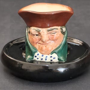 以下が含まれることがあります： 茶色の三角帽子と白と青の水玉模様のクラバットが付いた緑色のコートを着た人物を模した陶器製のキャラクタージャグ。このフィギュアは、光沢のある黒い円形の台座の中央に配置されており、装飾的なテーブルトップの置物として構成されています。