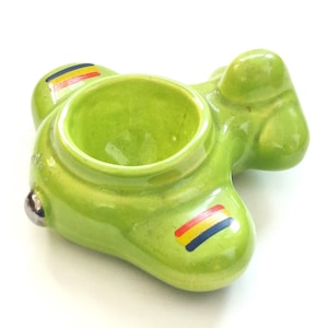 May include: A lime green ceramic egg cup shaped like a small airplane. The body features a central circular opening, a silver-toned nose piece, and wings decorated with horizontal stripes in red, yellow, and blue colors on a glossy glazed surface.