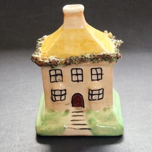 May include: A ceramic house figurine featuring a cream-colored facade with black painted windows and a dark red door. The structure has a yellow roof with textured green foliage along the eaves and sits on a green base with painted steps.