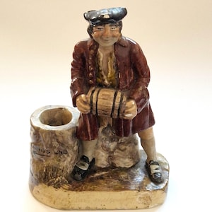 Può includere: Una statuetta in ceramica di una persona che indossa un cappotto rosso scuro e un cappello nero, che tiene in mano un piccolo barile di legno. La figura è seduta accanto a un ceppo cilindrico cavo con la scritta LANDS END sul lato.