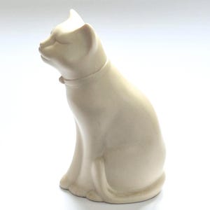 May include: A smooth, matte off-white ceramic cat figurine in a seated position. The cat is depicted with its head turned slightly upward and to the left, featuring a simple collar around its neck and a long tail curled neatly at its base.