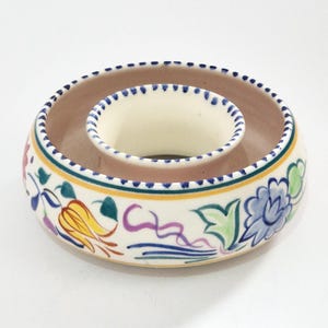 Può includere: Una ciotola rotonda in ceramica con un anello centrale rialzato, caratterizzata da un motivo floreale dipinto a mano in giallo, blu, verde e viola. Il bordo è decorato con una linea tratteggiata blu e l'interno ha una finitura beige chiaro.