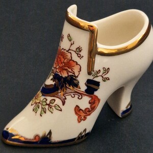 May include: A white porcelain shoe-shaped figurine featuring a floral pattern in orange, blue, and green. The decorative object has a gold-painted trim along the top edge, the toe, and the heel, with a glossy finish and a hollow interior opening.