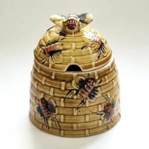 May include: A ceramic honey pot shaped like a woven beehive with a glossy tan finish. The lid features a sculpted bee handle, and the body is decorated with raised, hand-painted bees in brown and black tones, featuring a small cutout for a spoon.