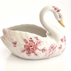 Könnte beinhalten: Eine weiße Keramikschale in Schwanenform mit handgemalten rosa Blumenmustern auf Körper und Flügeln. Der Schwan hat einen langen, geschwungenen Hals, einen gelben Schnabel und eine hohle Mitte, akzentuiert durch zarte rosa Blattdetails am Kopf.