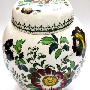May include: A round ceramic ginger jar with a matching lid, featuring a white base decorated with colorful floral patterns in shades of red, yellow, green, and dark purple. The lid is adorned with a green scalloped border and floral motifs.