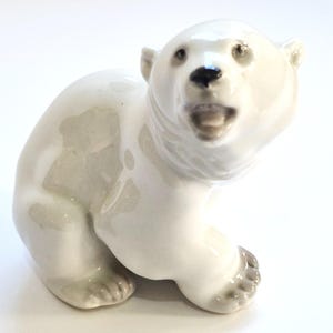 Puede incluir: Una figura de porcelana blanca brillante de un cachorro de oso polar en posición sentada. El cachorro tiene un acabado liso y reflectante, ojos pintados oscuros, una nariz negra y la boca abierta mostrando los dientes.