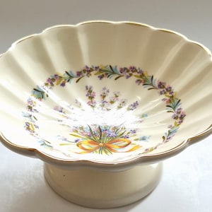 May include: A cream-coloured ceramic pedestal bowl with a scalloped edge and a gold-trimmed rim. The interior features a floral pattern of purple and blue flowers tied with an orange ribbon, arranged in a circular wreath design on the base.
