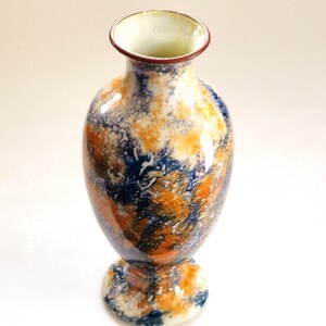 May include: A ceramic vase with a mottled, multi-coloured glaze featuring swirling patterns of deep navy blue, vibrant orange, and creamy white. The vessel has a rounded body, a narrow neck, a flared rim with a dark brown edge, and a circular base.