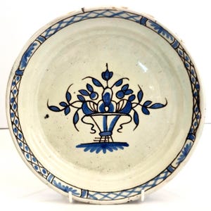 Könnte beinhalten: Ein runder Keramikteller mit cremeweißer Glasur und einem handgemalten blauen Blumenmotiv in der Mitte. Das Design zeigt einen stilisierten Korb gefüllt mit Blumen und Blättern. Der Rand ist mit einem blauen Rauten- und Blattmuster verziert.