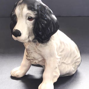 以下が含まれることがあります： 白い体に頭と耳に黒い斑点がある、座っている子犬の陶器製フィギュア。子犬は黒い鼻、茶色の目をしており、滑らかで光沢のある仕上がりです。この彫刻は、前足を前に伸ばして座っている犬の姿を捉えており、詳細に成形された毛並みの質感とリアルな顔立ちが特徴です。