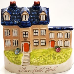 Peut inclure: Une figurine en céramique de Stanfield Hall représentant un bâtiment beige avec un toit bleu, deux portes orange et plusieurs fenêtres. La base est peinte en vert avec de petites fleurs, et le texte Stanfield Hall est inscrit sur le devant.