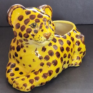 May include: A ceramic planter shaped like a reclining leopard with a bright yellow base coat and dark brown spots. The leopard has a smooth, glossy finish, pointed ears, and a hollow opening on its back for plants. The interior is cream colored.