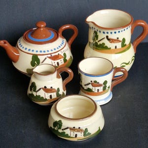 Op de afbeelding: Een crèmekleurige keramische theeset met bruine accenten. De set bevat een theepot, een melkkan, een suikerpot en een mok. Elk stuk is versierd met een geschilderde huis- en boomscène. De theepot heeft een bruin deksel en handvat.