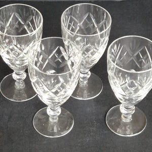 May include: Four clear crystal wine glasses with diamond-cut patterns. Each glass has a round base, a stem, and a bowl. The glasses are arranged on a dark surface.