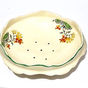 Może przedstawiać: Kremowa ceramiczna miseczka na owoce jagodowe z falistym brzegiem i zielonym wykończeniem. Wnętrze zdobi ręcznie malowany wzór zielonych liści oraz kiści żółtych i pomarańczowych owoców po obu stronach, z siedmioma otworami drenażowymi w dnie.