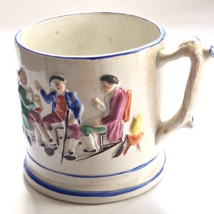 May include: A cream-coloured ceramic mug featuring a raised, hand-painted relief scene of figures in historical attire. The mug has a blue rim and base trim, and a handle shaped like a textured tree branch. The exterior displays colourful painted details.