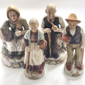 Może przedstawiać: Zestaw czterech porcelanowych figurek przedstawiających starsze osoby w wiejskich strojach. Dwie postacie siedzą na okrągłych podstawach, a dwie stoją. Ubrane są w stonowane odcienie brązu, zieleni i bieli, z akcesoriami takimi jak kosze, laski i fajka.
