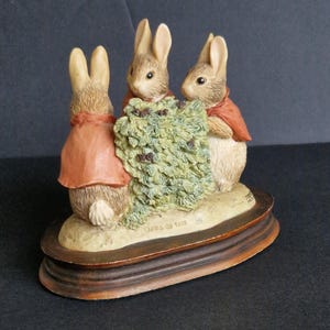 Può includere: Statuetta in ceramica raffigurante tre conigli con mantelli rossi riuniti attorno a un cespuglio verde. I conigli hanno la pelliccia marrone chiaro e sono posizionati su una base color crema con un bordo marrone a più livelli. Oggetto da collezione.