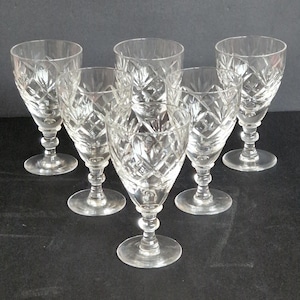 Pode incluir: Seis taças de vinho de cristal transparente com padrões lapidados em diamante. As taças têm um design clássico com base redonda, haste e taça. As taças estão dispostas sobre uma superfície escura.