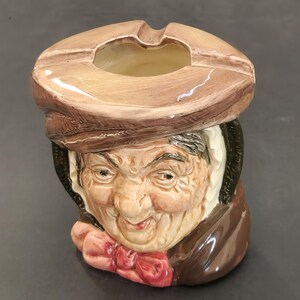 以下が含まれることがあります： 茶色の帽子とピンクの蝶ネクタイを身につけた人物の形をした陶器製の灰皿。顔の造形は細かく、しわがはっきりと見え、光沢のある仕上げが施されている。帽子の頂部には、灰を捨てるための円形の開口部がある。