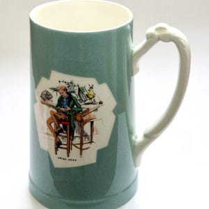 Puede incluir: Una taza de cerámica de color verde claro con interior blanco y un asa decorativa. El frente presenta una ilustración impresa de una persona sentada en un taburete frente a un escritorio, con el texto "URIAH HEEP" en letras negras debajo.