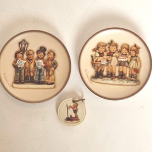 Puede incluir: Tres platos decorativos y una pequeña caja de música redonda con figuras de Hummel. Los platos tienen un fondo beige con bordes marrones y representan a niños cantando y tocando instrumentos. La caja de música tiene una pequeña figura de un niño tocando un instrumento.