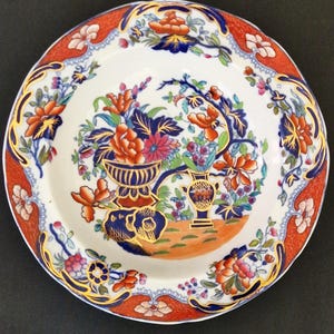 May include: A round, white ceramic plate with a vibrant floral design. The plate features a red and orange border with floral accents. The center showcases a detailed arrangement of flowers in vases, with blue, orange, and pink hues, accented with gold.