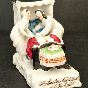 May include: A white porcelain figurine showing two figures in white robes by a bed. The bed has a red blanket and a green and white patterned pillow. The base of the figurine has the inscription "The last to put out the light."