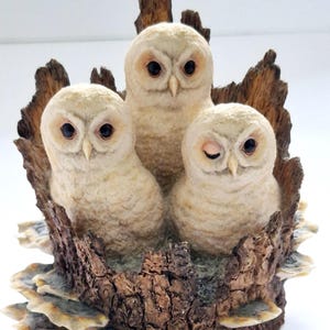 May include: A decorative figurine featuring three cream-colored owls nestled inside a hollowed-out tree trunk. The owls have dark eyes, with the owl on the right appearing to wink. The tree trunk has a textured brown bark finish.