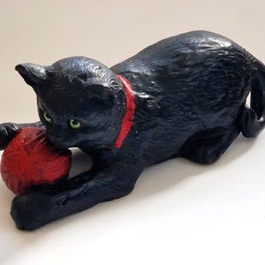 May include: A black cat figurine in a crouching position with green eyes and a red collar. The cat is playing with a textured red ball of yarn held between its front paws. The sculpture has a detailed, molded surface texture throughout its body.