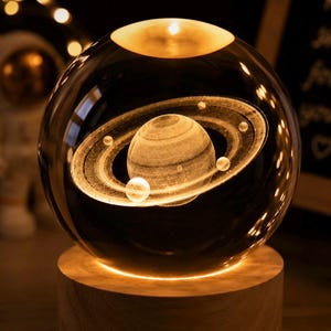 May include: A clear glass sphere containing a three-dimensional laser-etched model of the planet Saturn with its rings and several moons. The sphere is illuminated from below and rests on a cylindrical light-coloured wooden base.