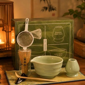 Peut inclure: Un ensemble de thé matcha avec un bol en céramique vert, un fouet, un tamis et une cuillère. L'ensemble comprend un support à fouet et est présenté sur un tapis décoratif. La boîte contient des illustrations et du texte.