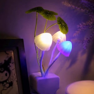 May include: A mushroom-shaped night light featuring three illuminated caps in white, soft yellow, and blue-purple hues. The lamp has thin green stems and realistic green leaves extending from a solid, rectangular white base that glows from within.
