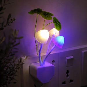 Op de afbeelding: Een decoratief nachtlampje in de vorm van paddenstoelen, met wit en blauw oplichtende doppen en groene bladeren. Het nachtlampje wordt in een stopcontact gestoken en geeft een zachte, omgevingsgloed, perfect voor een slaapkamer of kinderkamer.