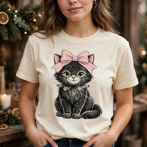 Può includere: Una t-shirt a maniche corte color crema con un'illustrazione grafica di un gattino nero dagli occhi grandi, che indossa un fiocco rosa chiaro sulla testa e un colletto a balze intorno al collo, centrata sul davanti del capo.