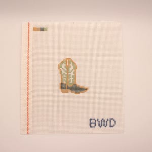 May include: A needlepoint canvas featuring a stitched design of a cowboy boot in shades of green, tan, and black. The boot is centered on a white canvas with the letters "BWD" stitched in blue.