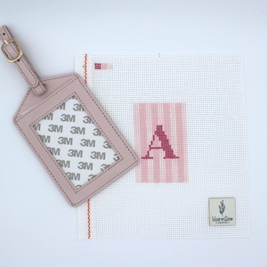 Puede incluir: Una etiqueta de equipaje rosa claro con una hebilla dorada junto a un lienzo de punto de aguja. El lienzo presenta un fondo a rayas rosas con la letra "A" cosida en un rosa más oscuro. Una pequeña etiqueta con el texto "blue willow" está en el lienzo.
