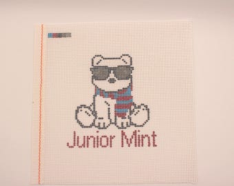 May include: Cross-stitch design featuring a bear wearing sunglasses and a scarf with red, blue, and white stripes. The text "Junior Mint" is stitched below the bear. The design is on a white canvas with a red border.
