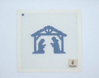 Op de afbeelding: Een vierkant, gebroken wit borduurcanvas met een blauwe kerststal. Het ontwerp toont een stal en twee figuren. Een klein, rechthoekig label met de tekst "blue willow designs" staat in de rechter benedenhoek.