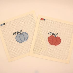 Op de afbeelding: Twee vierkante borduurkaders, één met een blauw pompoenontwerp en de andere met een rood pompoenontwerp. Elke pompoen is gestikt met een zwarte stengel en blad details. De achtergrond is crèmekleurig.