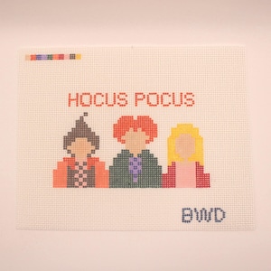 Könnte beinhalten: Ein Kreuzstich-Design auf weißer Leinwand mit dem Text "HOCUS POCUS" in Rot. Darunter befinden sich drei pixelige Figuren mit unterschiedlichen Haar- und Kleiderfarben. Die Initialen "BWD" sind blau gestickt.